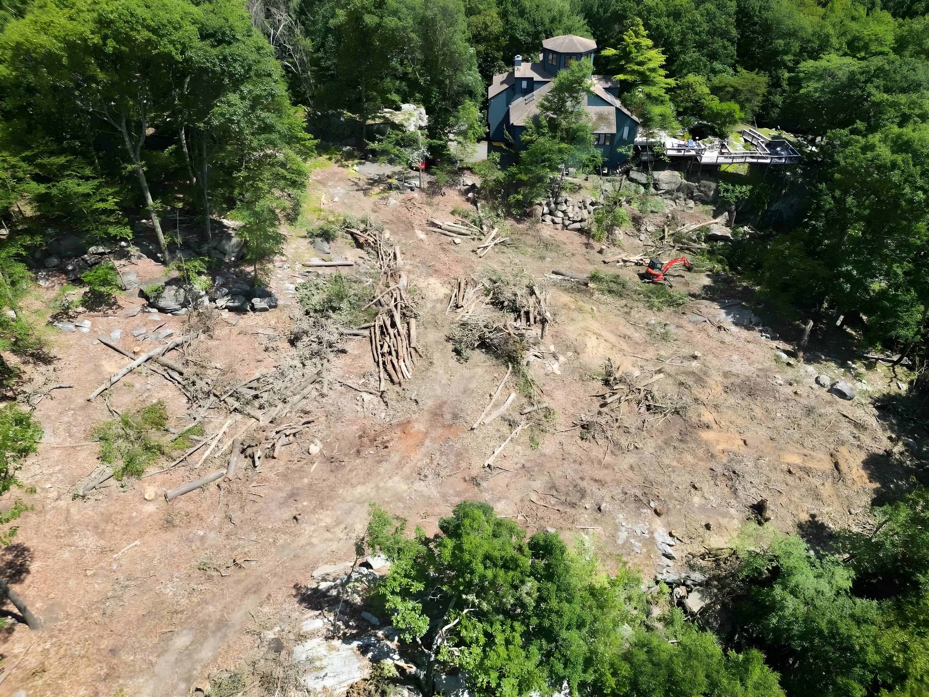 Drone aerial of a cleared hillside with staged log piles and a mini excavator at work