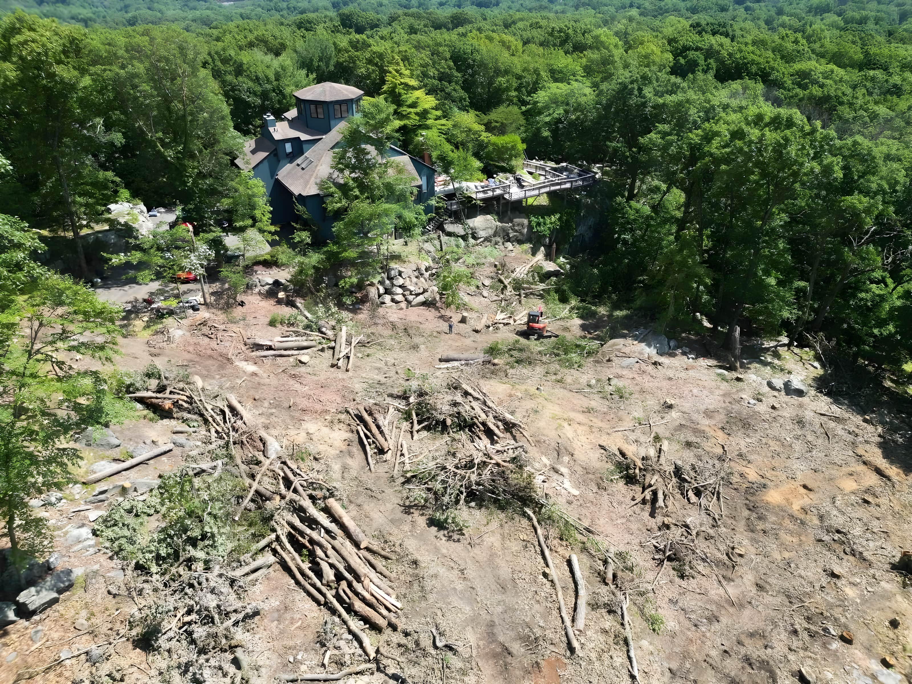 Drone aerial of a rocky hillside lot cleared of trees with log piles and a mini excavator