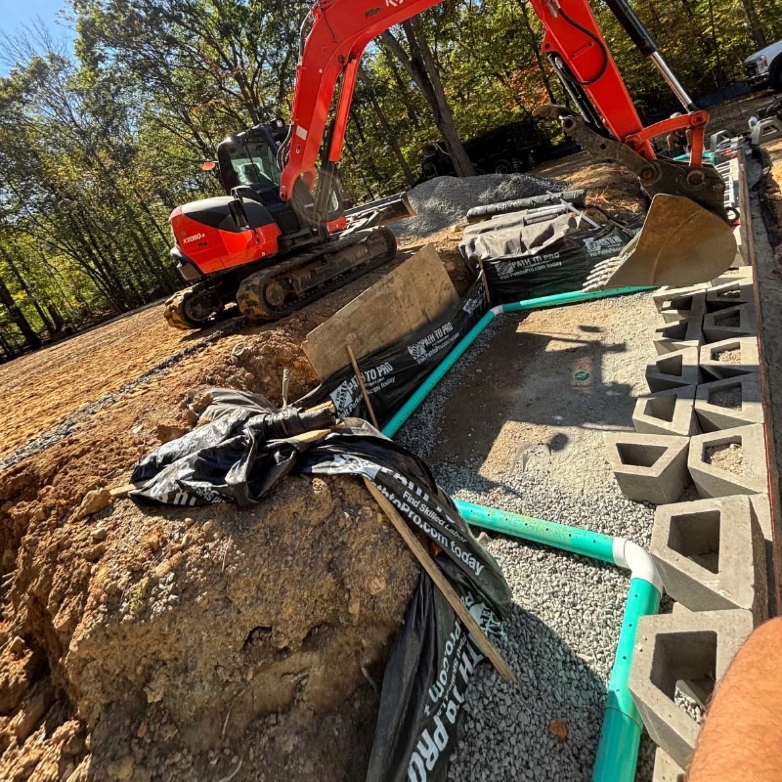 Excavator beside a gravel lined foundation drain with PVC pipe, filter fabric, and wall block